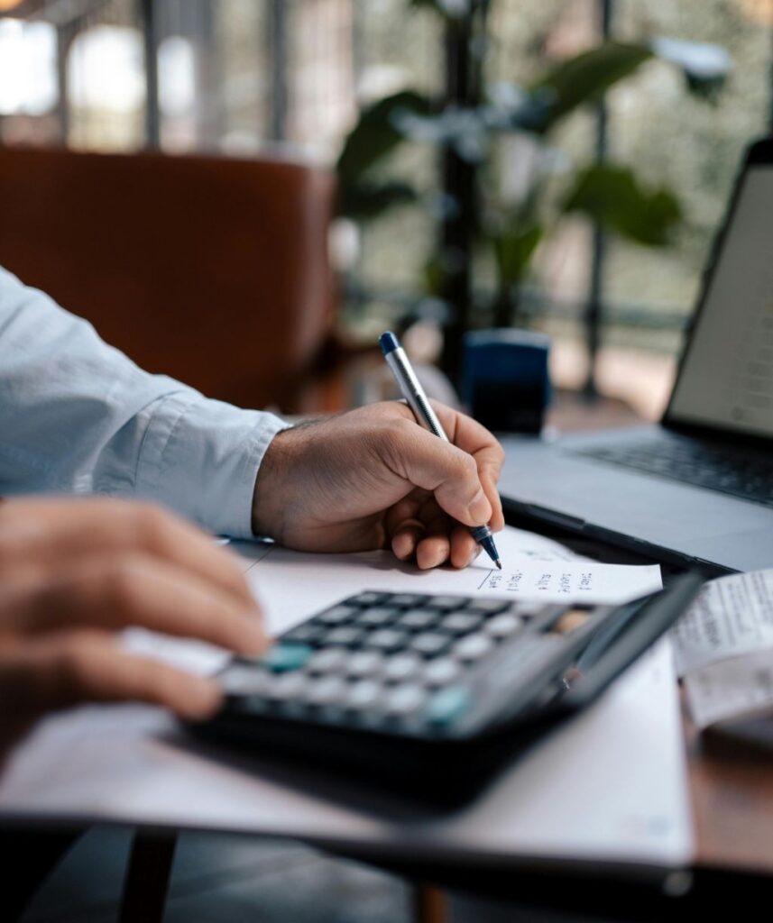 Main d’un collaborateur utilisant une calculatrice et prenant des notes sur un bureau, travail comptable en environnement professionnel.