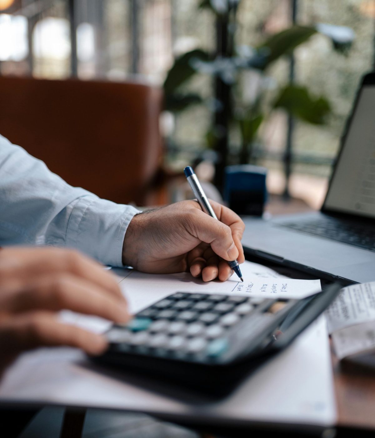 Main d’un collaborateur utilisant une calculatrice et prenant des notes sur un bureau, travail comptable en environnement professionnel.